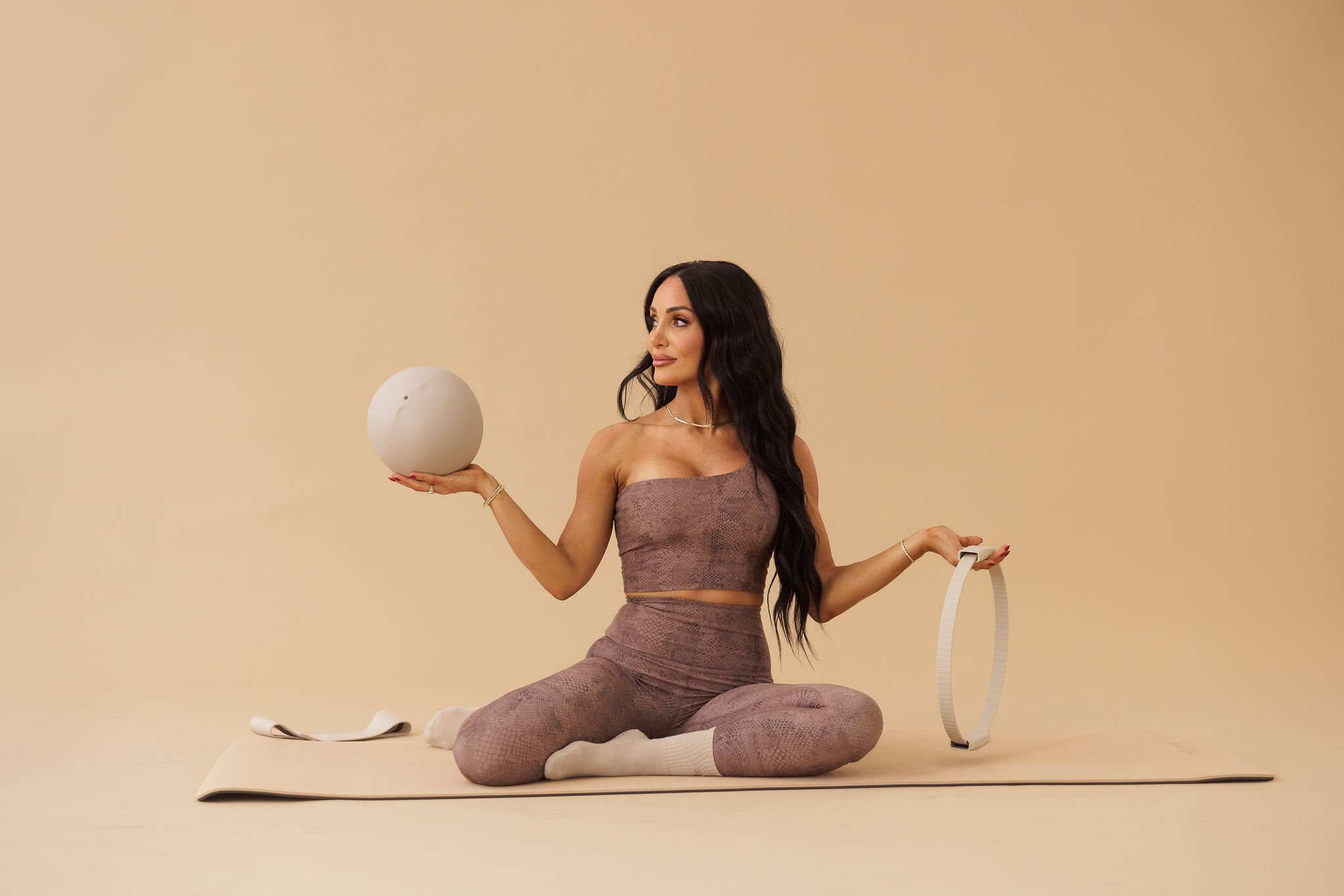 Pilates instructor seated on a mat holding a Pilates ball and ring against a neutral studio background wearing earth-tone activewear