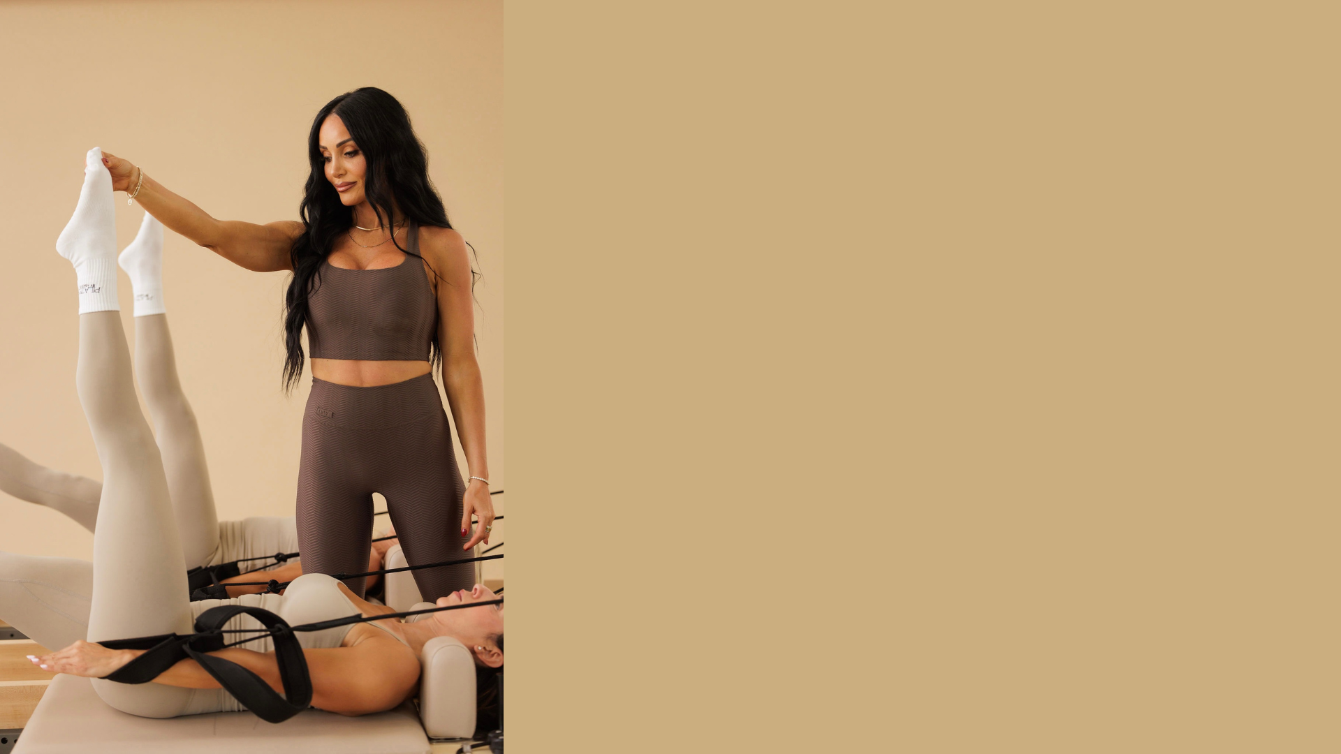 A Pilates instructor assists a client on a reformer, gently guiding extended legs to support proper alignment and controlled movement in a calm, neutral-toned studio setting.