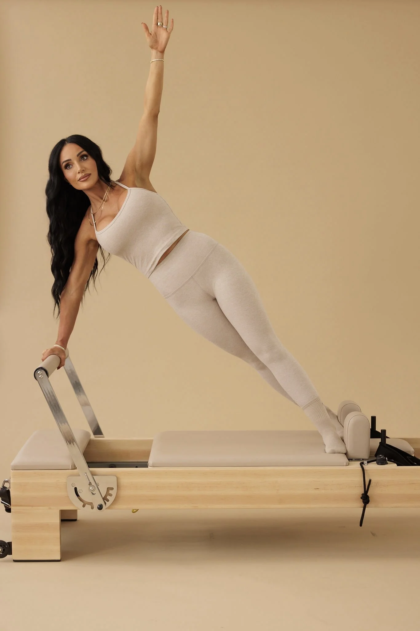 A Pilates instructor performing a controlled side plank variation using a reformer, emphasizing balance, strength, and fluid movement against a warm, neutral background.