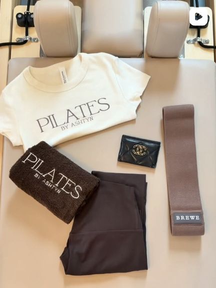Pilates branded apparel and studio essentials arranged neatly on a table, including a logo t shirt, towel, resistance band, and accessories