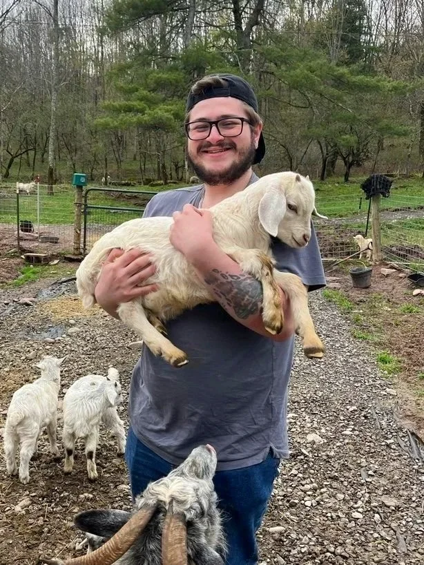 A man with glasses, a beard, and a black backwards cap holding a small light-colored goat, smiling outdoors on a farm with trees and other goats in the background.