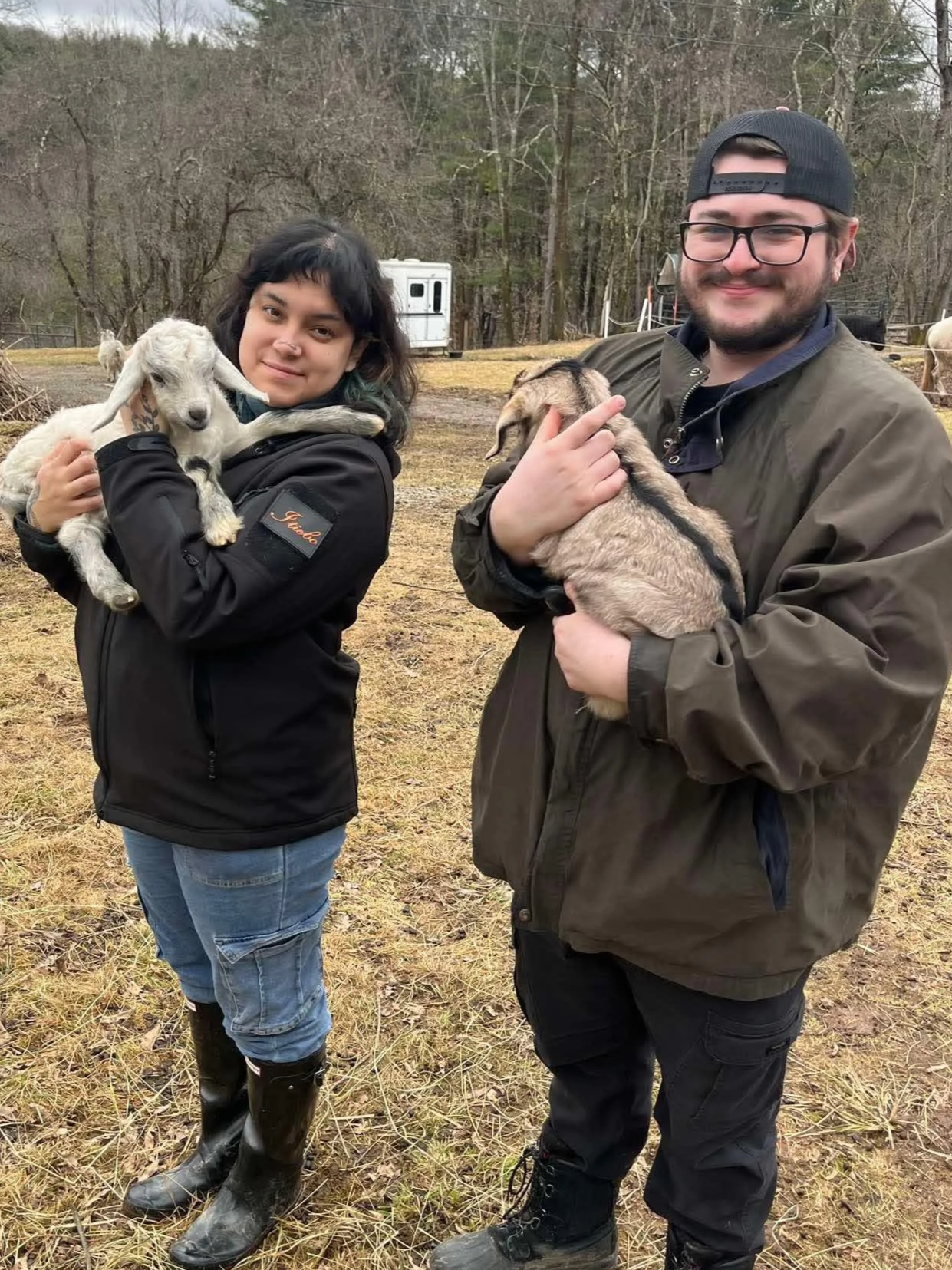 Two people holding baby goats outdoors in a farm or rural setting, with trees and a trailer in the background.