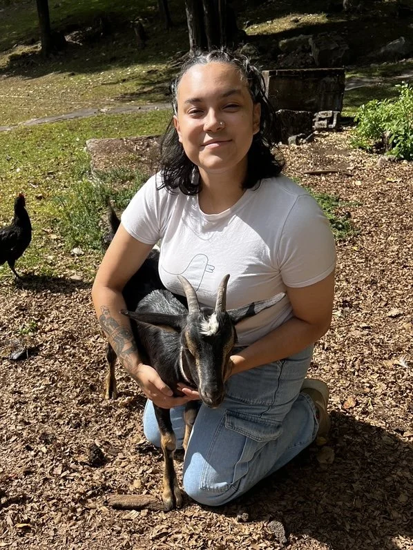 A young woman with dark curly hair and a tattoo on her arm, kneeling on the ground, holding a small goat. She is smiling and outdoors in a wooded area with sunlight filtering through the trees.
