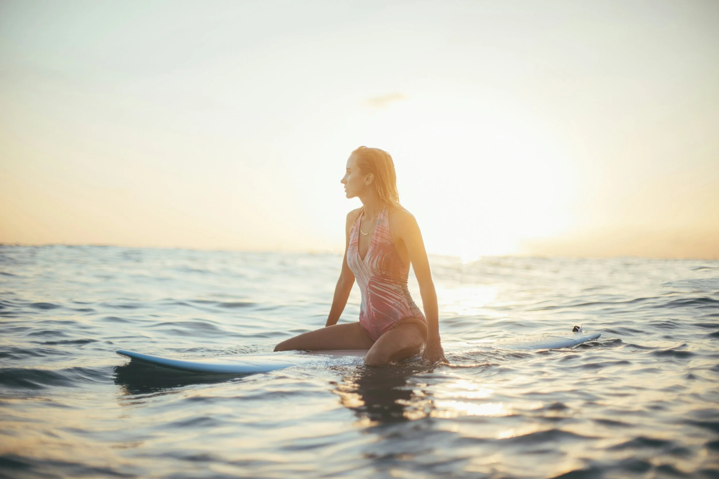 Image of a woman sitting on a surfboard in the ocean at dawn, displaying a fit physique while she waits for a wave.