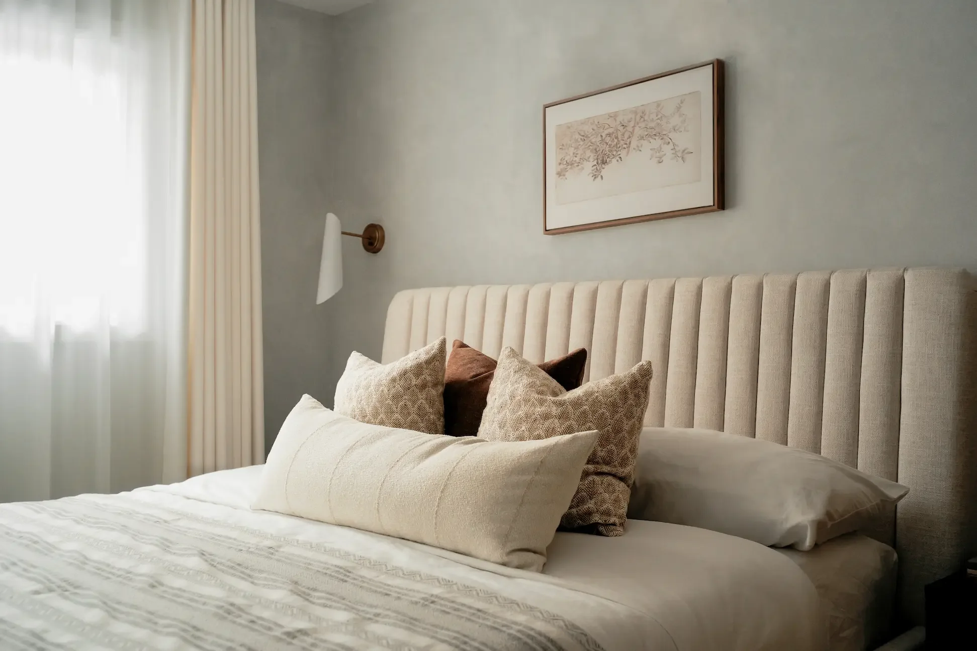 Serene modern bedroom in Toronto by Claire Design Studio, featuring a beige channel-tufted headboard, curated textured pillows, a minimalist brass wall sconce, and elegant wall art.