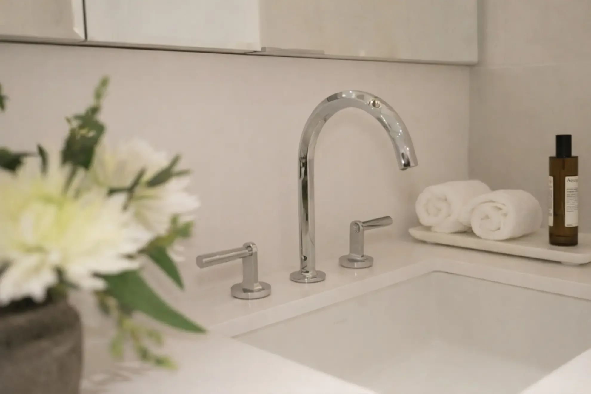 Close-up of a luxury bathroom vanity in Toronto by Claire Design Studio, featuring a polished chrome faucet, white stone countertop, and professional styling with fresh white flowers and rolled towels.