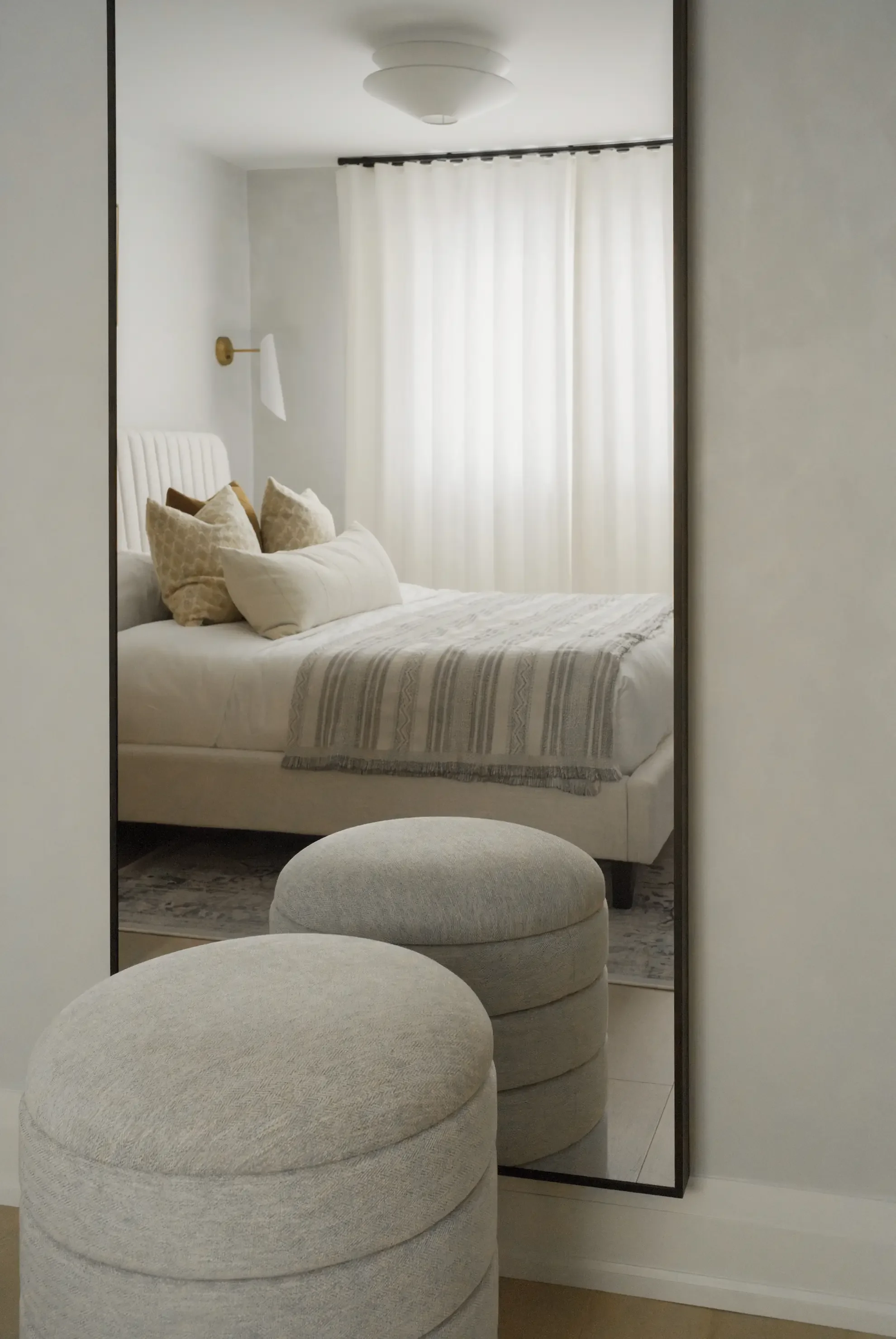 Modern transitional bedroom in Toronto by Claire Design Studio, captured through a tall floor mirror reflecting a neutral-toned bed with textured pillows, sheer white curtains, and two soft grey round poufs in the foreground.