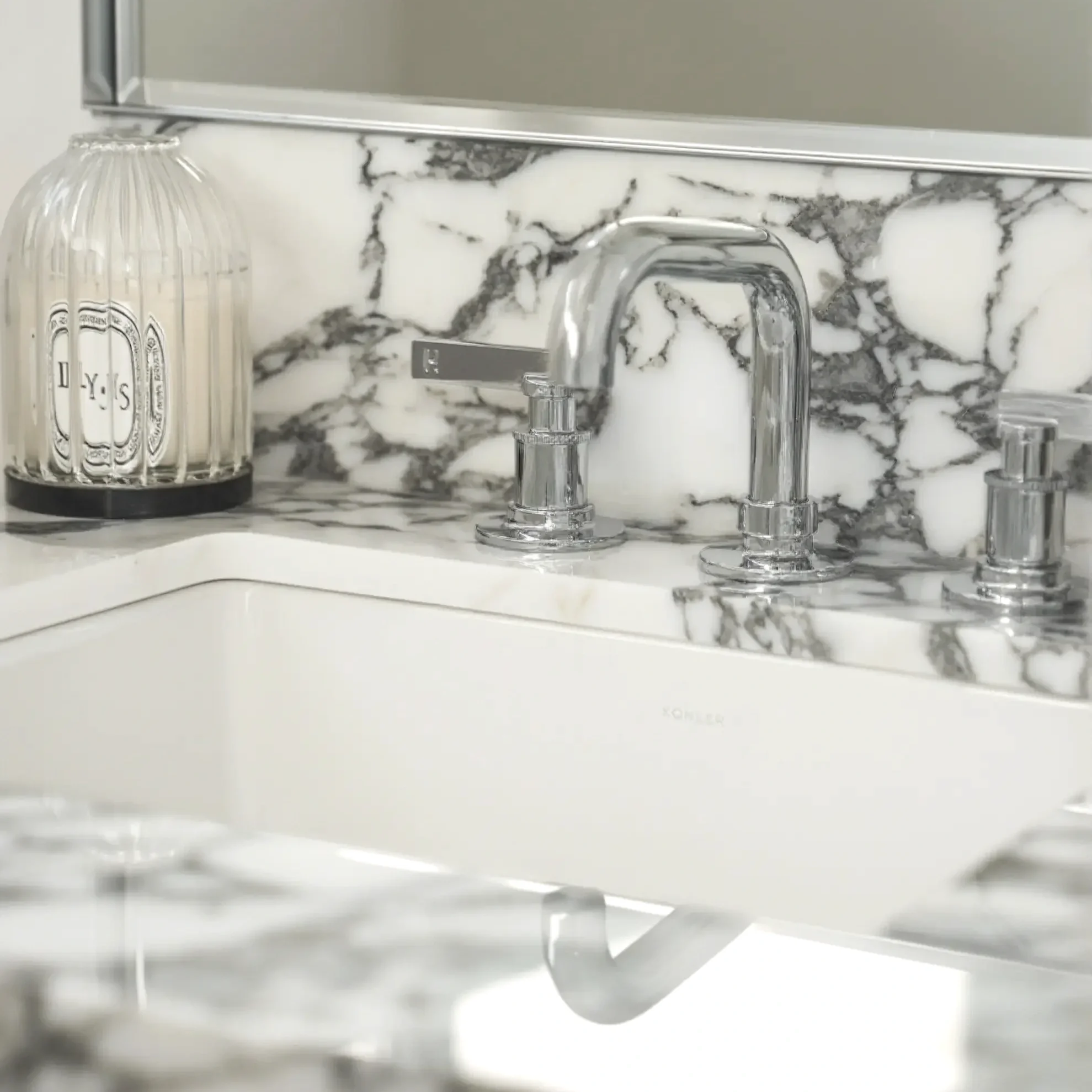 Close-up of a luxury bathroom vanity featuring bold Arabescato marble countertop, a polished chrome faucet, and a white Kohler undermount sink styled with a Diptyque candle.
