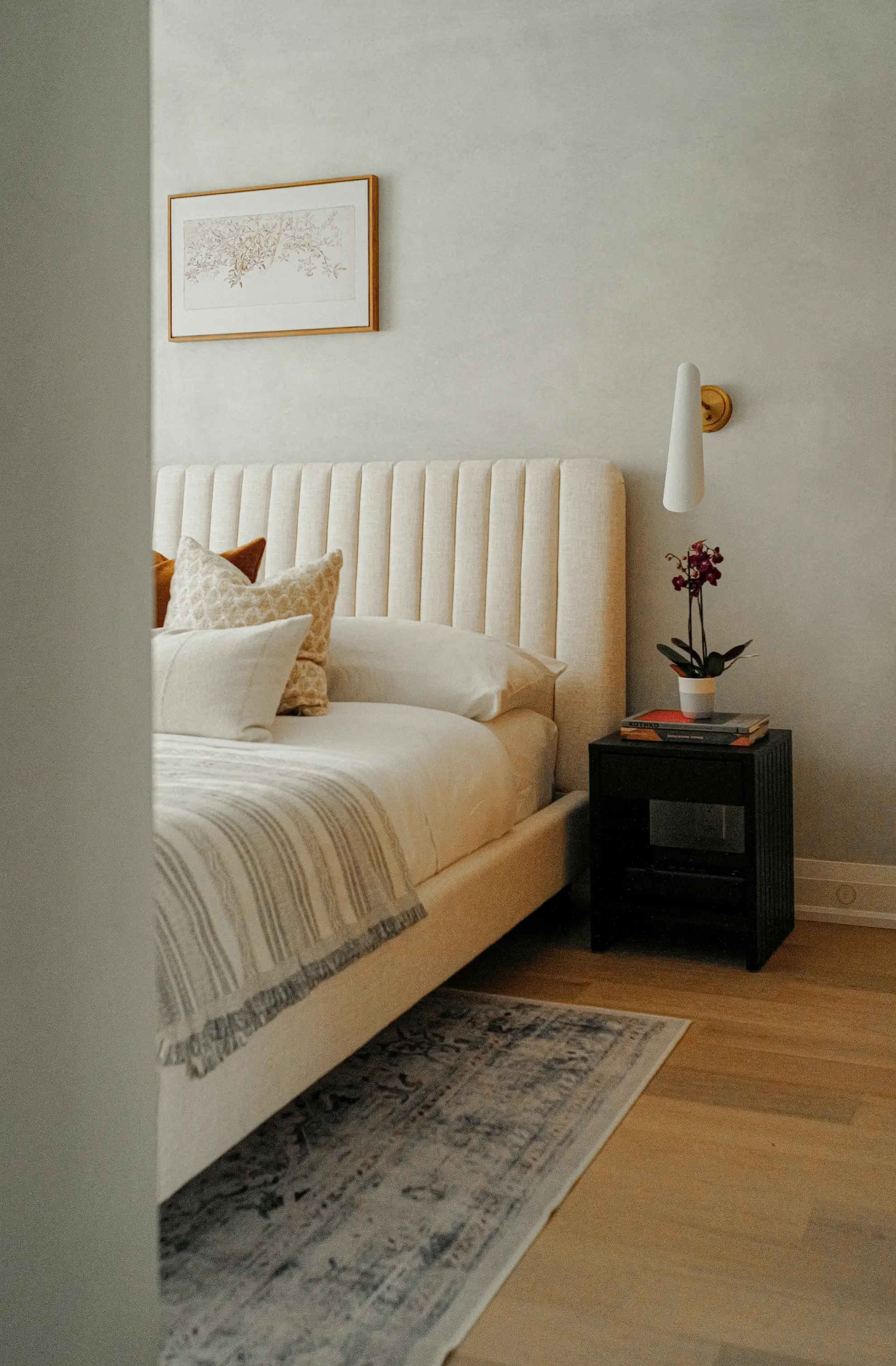 Modern bedroom detail by Claire Design Studio in Toronto, featuring a custom channel-tufted headboard, a black textured nightstand styled with a small orchid, and a brass wall sconce.