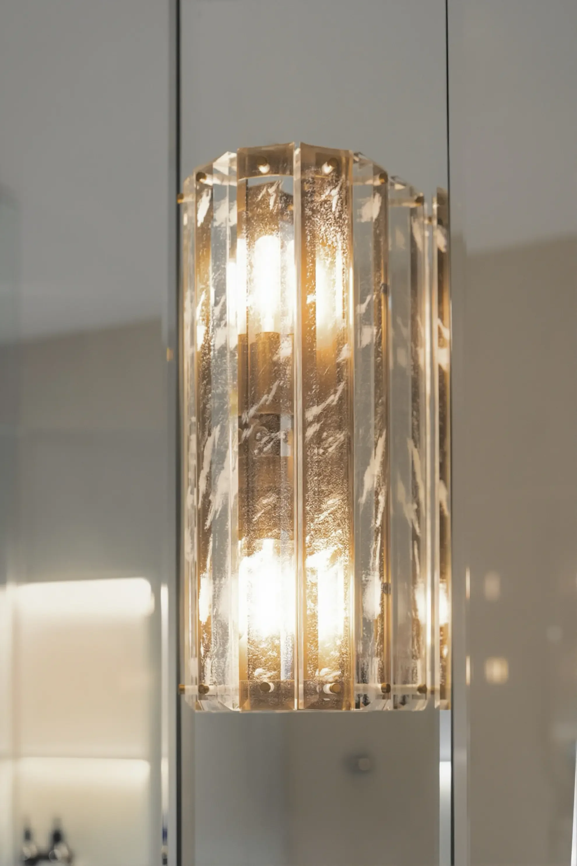 Detailed shot of an elegant pillar-style crystal wall sconce with gold accents, reflecting in a mirrored surface in a luxury Toronto bathroom designed by Claire Design Studio.