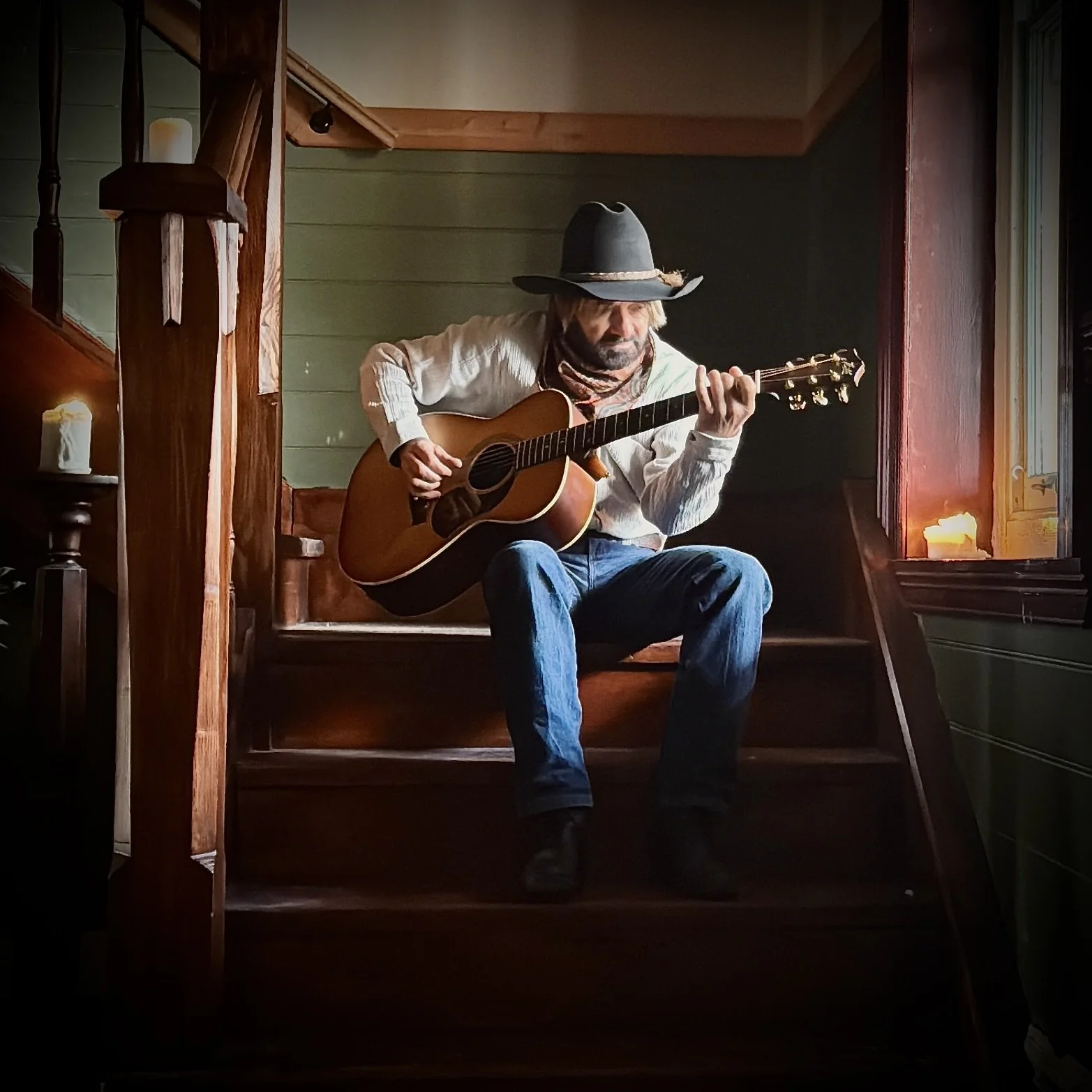 John Stokes - Serenading cowboy, white shirt, and jeans playing an acoustic guitar while sitting on wooden stairs near a window with candles.