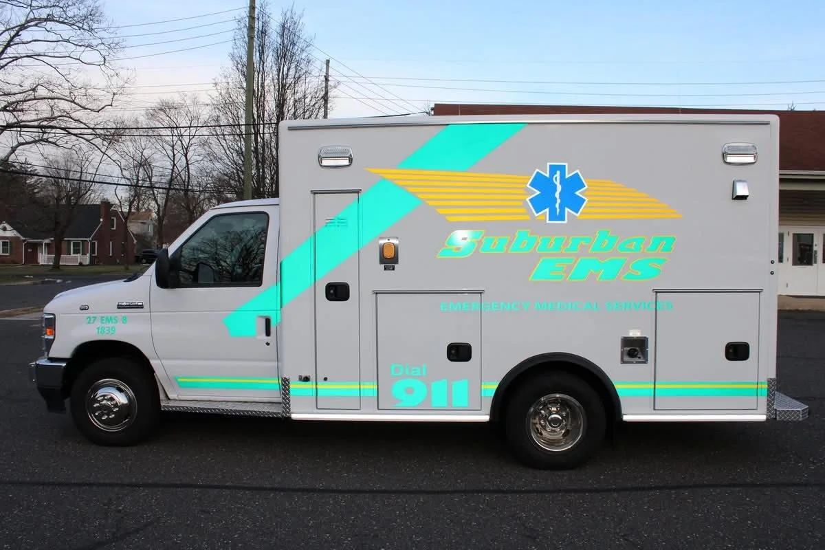 Side view of a white ambulance vehicle with Suburban EMS branding, emergency medical services text, a blue medical symbol, and a large luminous teal and yellow arrow design.