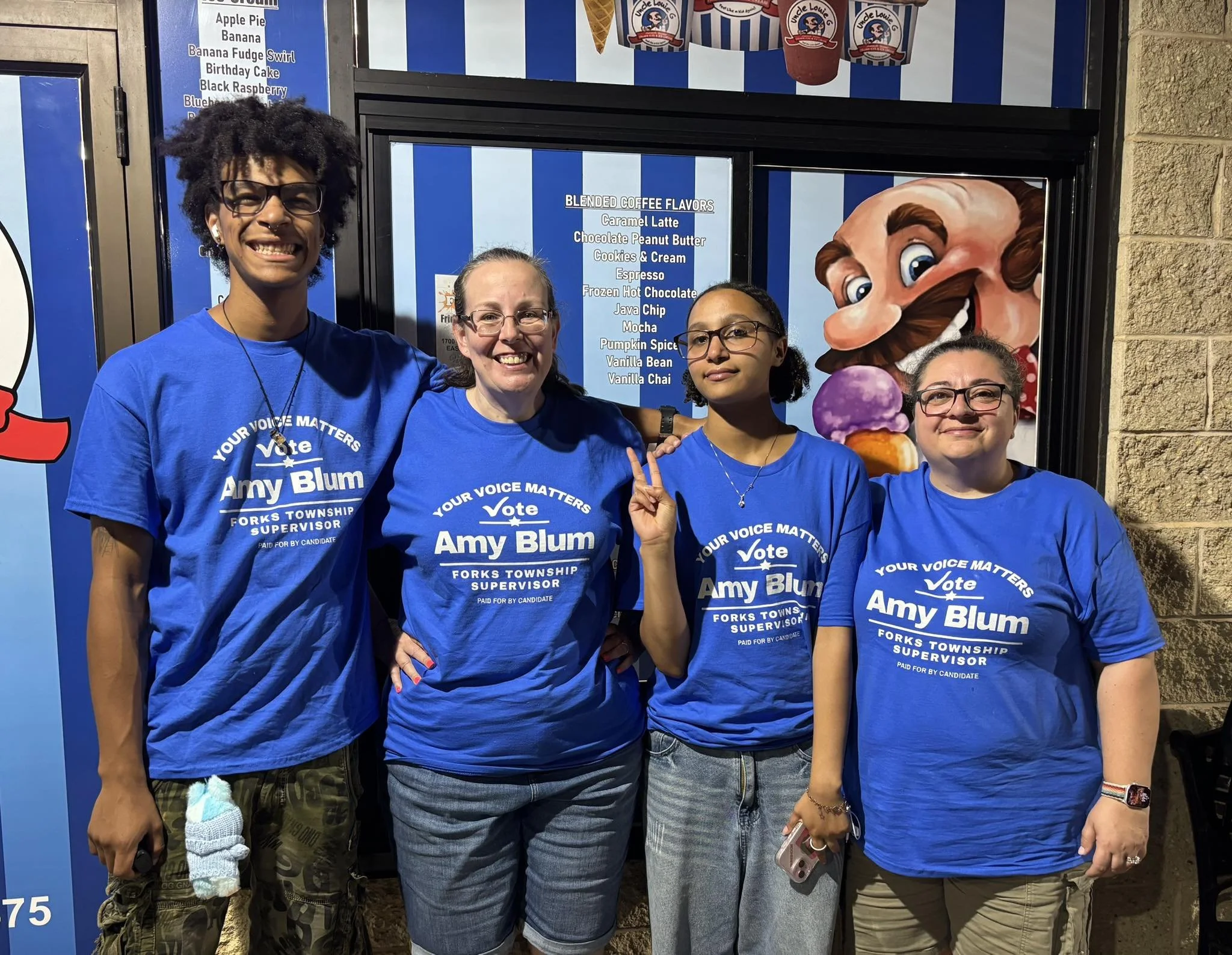 Four women standing outside an ice cream shop, wearing matching blue campaign shirts that read 'Your Voice Matters, Vote Amy Blum, Forks Township Supervisor, Paid for by Candidate.' The women are smiling or posing, with one flashing a peace sign, and there is colorful ice cream and a cartoon character in the background.