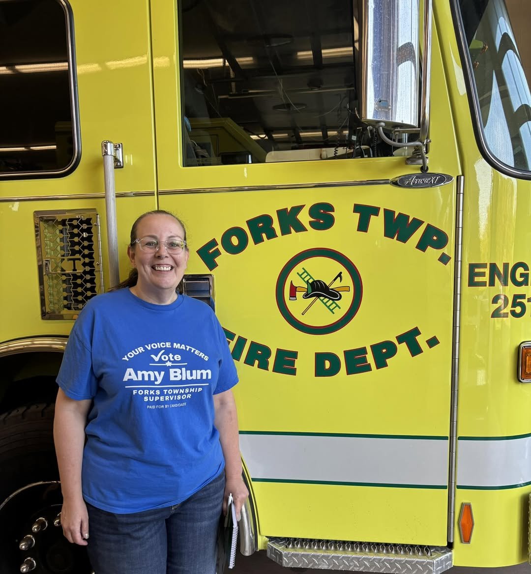 Amy Blum smiling and holding a folder, standing in front of a yellow fire truck with 'FORKS TWP FIRE DEPT.' written on the side.