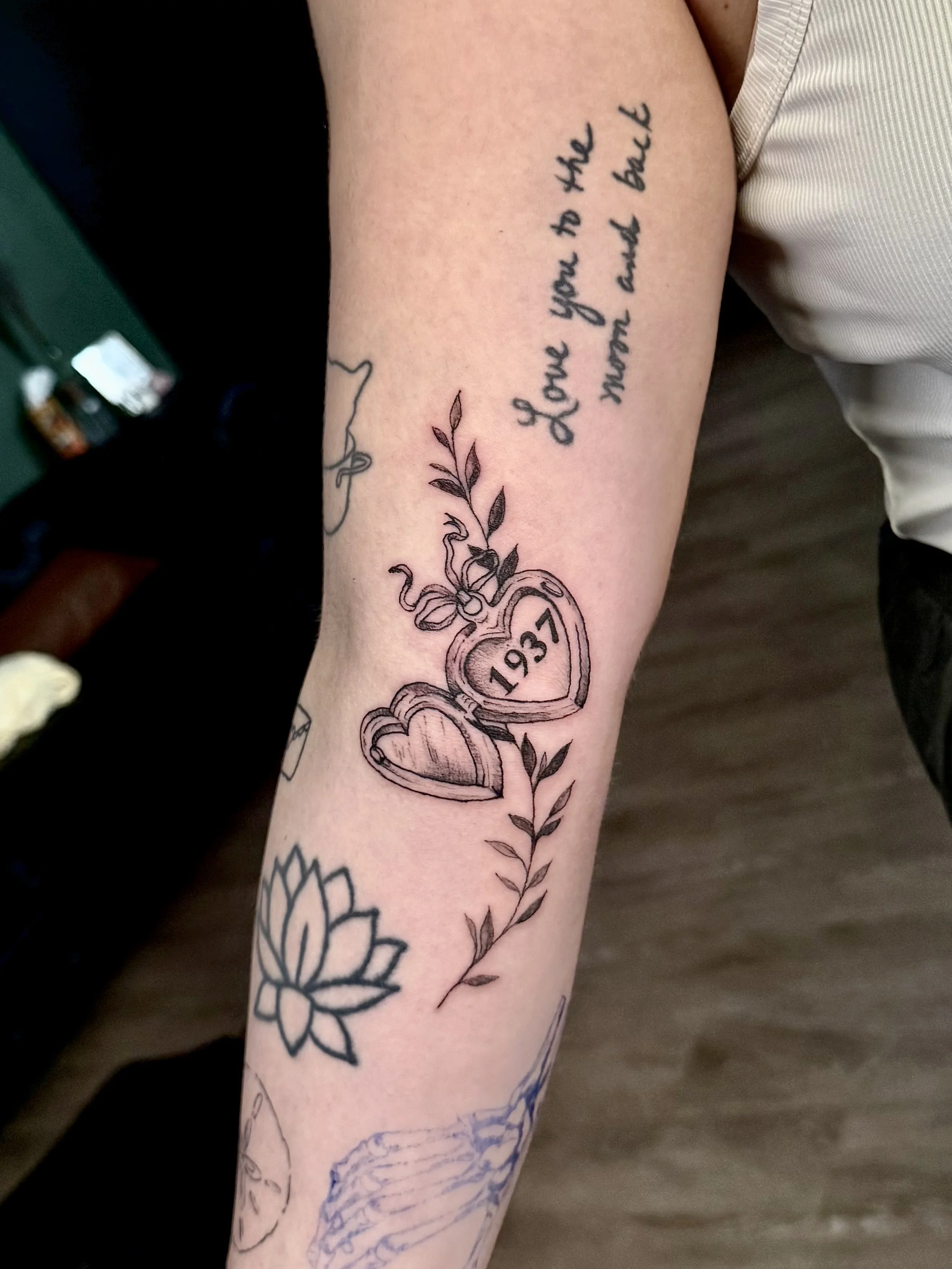 Tattoo of two intertwined hearts with the year 1937 inside one heart, surrounded by leaves. Above the tattoo, a handwritten-style text reads, "I love you to the moon and back."