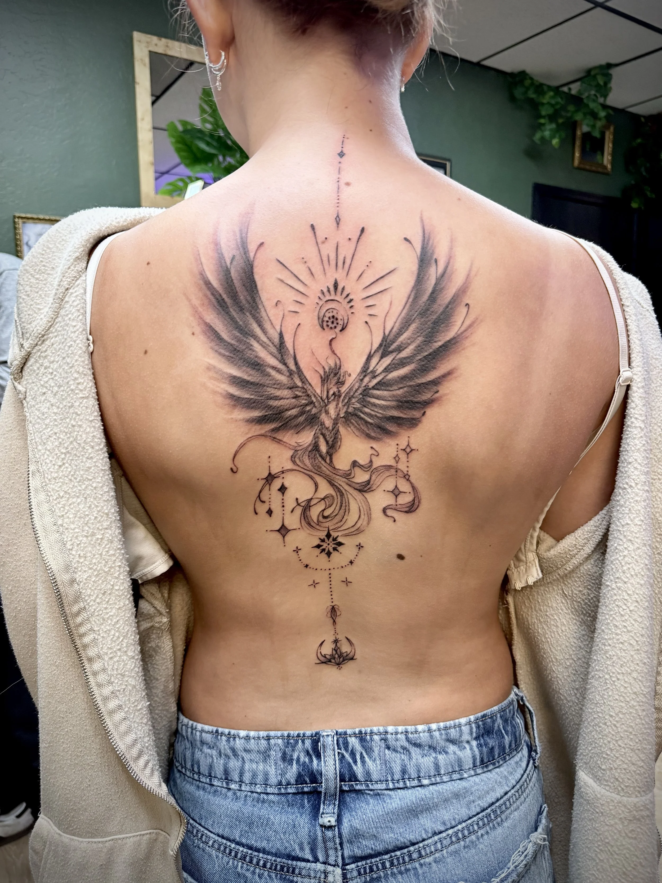 Back tattoo of a phoenix with outstretched wings, surrounded by celestial and geometric designs, with a sun and moon motif above and below.