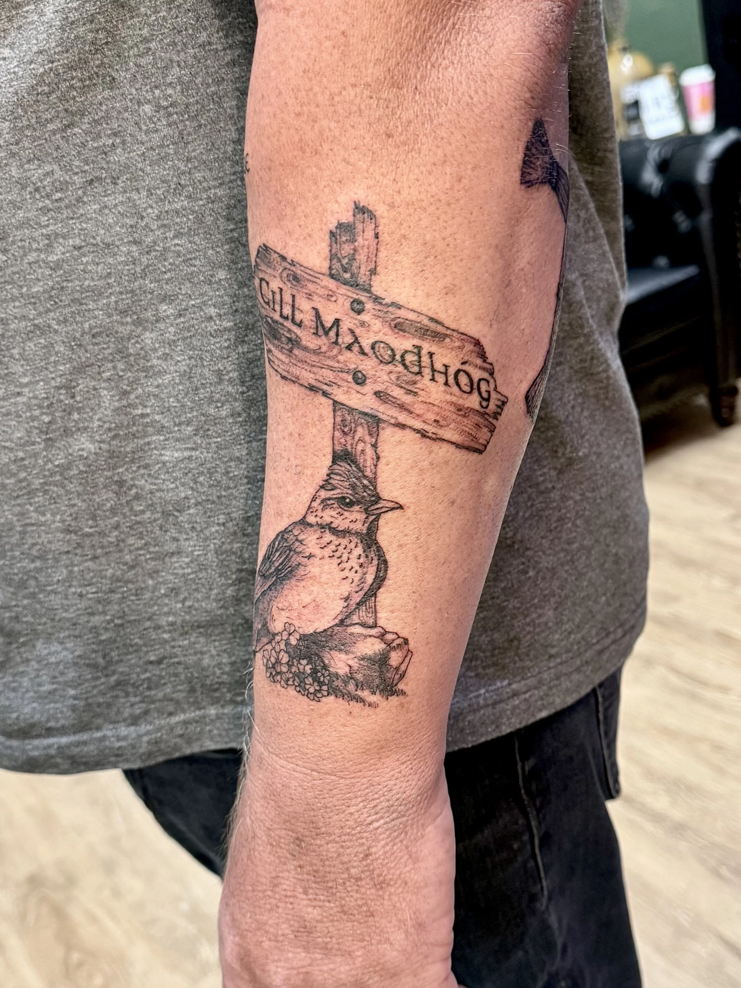 Tattoo of a bird sitting on a branch with a wooden sign hanging from the branch that reads "EAT BOWL GILL".