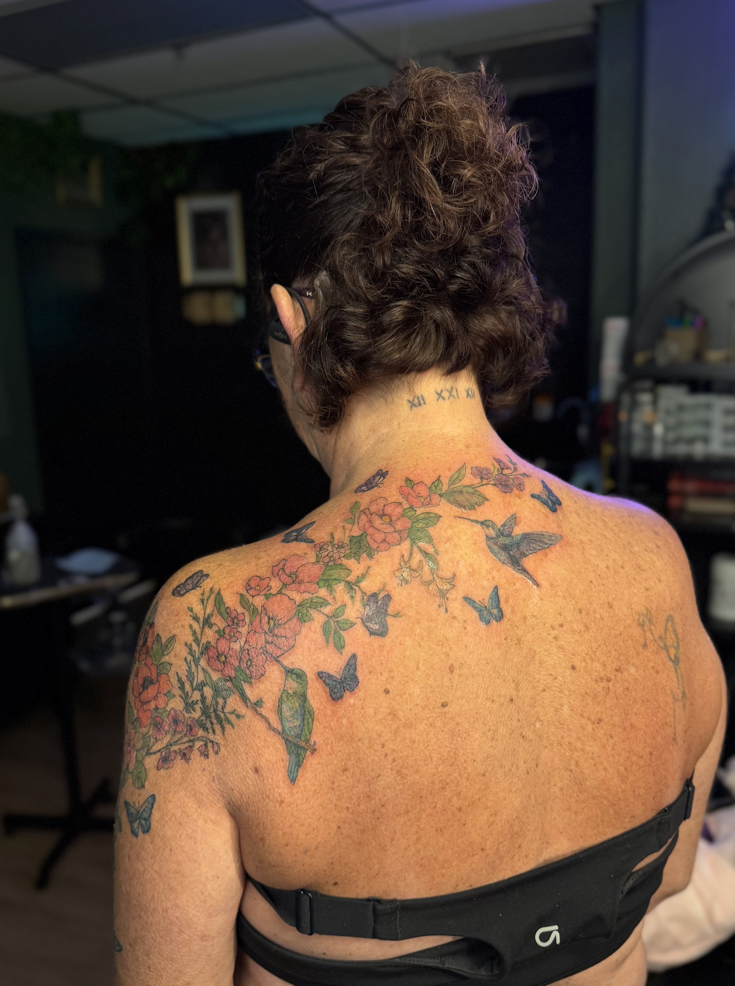 Back of a woman with a floral and butterfly tattoo across her shoulders and neck, wearing a black strapless top, in a room with a dark background.