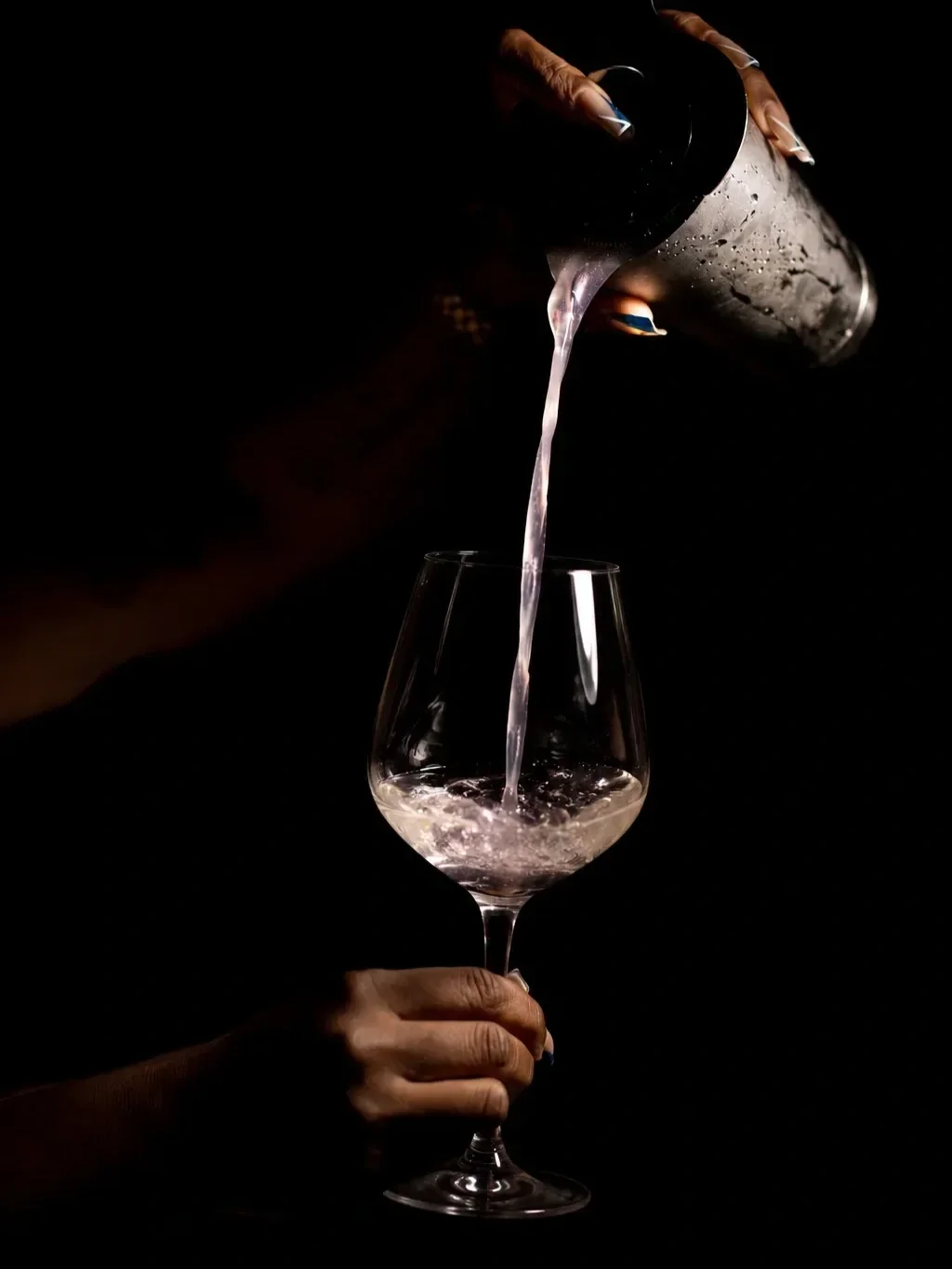 Person pouring water from a metal shaker into a wine glass against a black background.