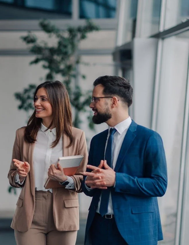 Two business professionals, a woman and a man, smiling and engaged in conversation in a modern office setting with large windows and greenery in the background.