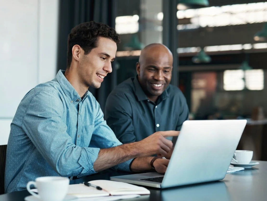 Two men sitting at a table in a modern office or café, looking at a laptop screen together, smiling and engaging in conversation.