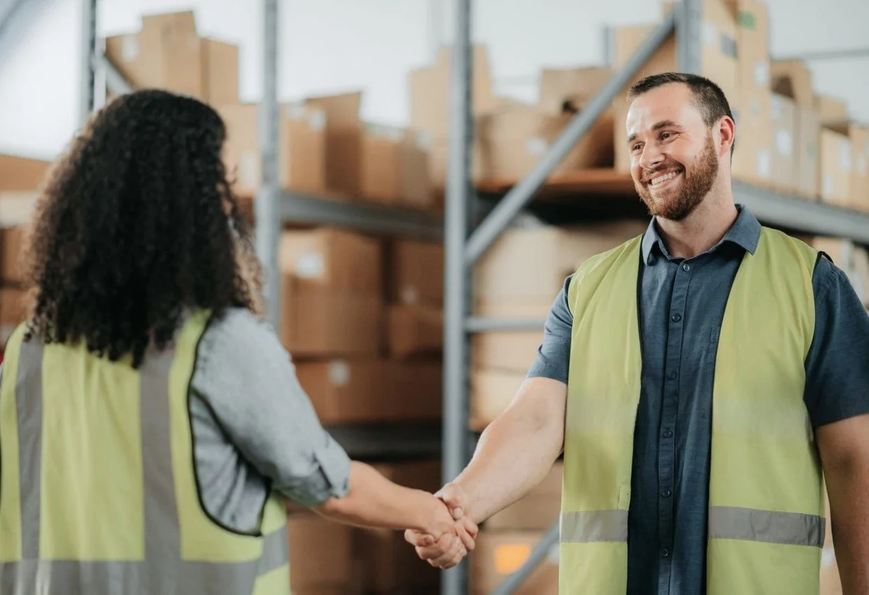 Two warehouse workers shake hands, smiling, with shelves of boxes in the background.
