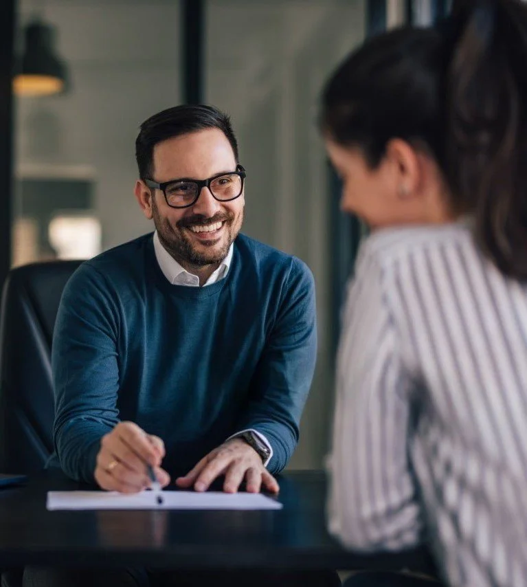 A man with glasses smiling and taking notes during a consultation with a woman in a striped shirt in an office setting.