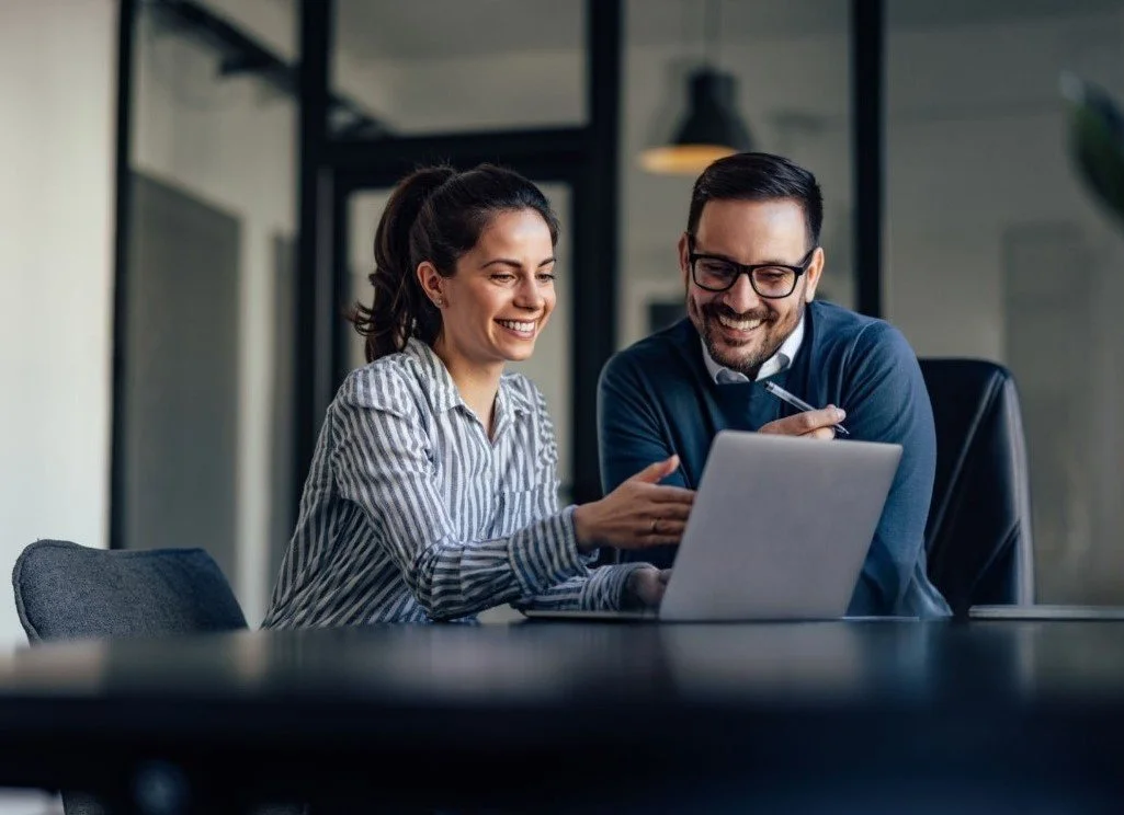 Two colleagues, a woman and a man, sitting at a desk looking at a laptop and smiling.