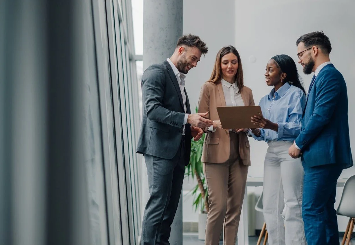 Four business professionals, two men and two women, are gathered around looking at a tablet in a modern office.