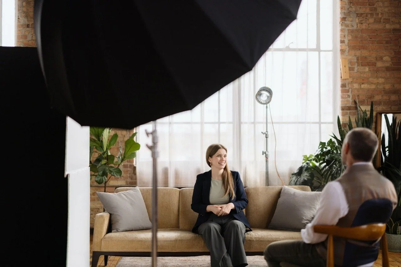 A woman seated on a beige sofa during an interview or conversation, smiling, with a man sitting opposite her in a room with large windows, brick wall, and plants, while a lighting umbrella and professional studio equipment are visible.