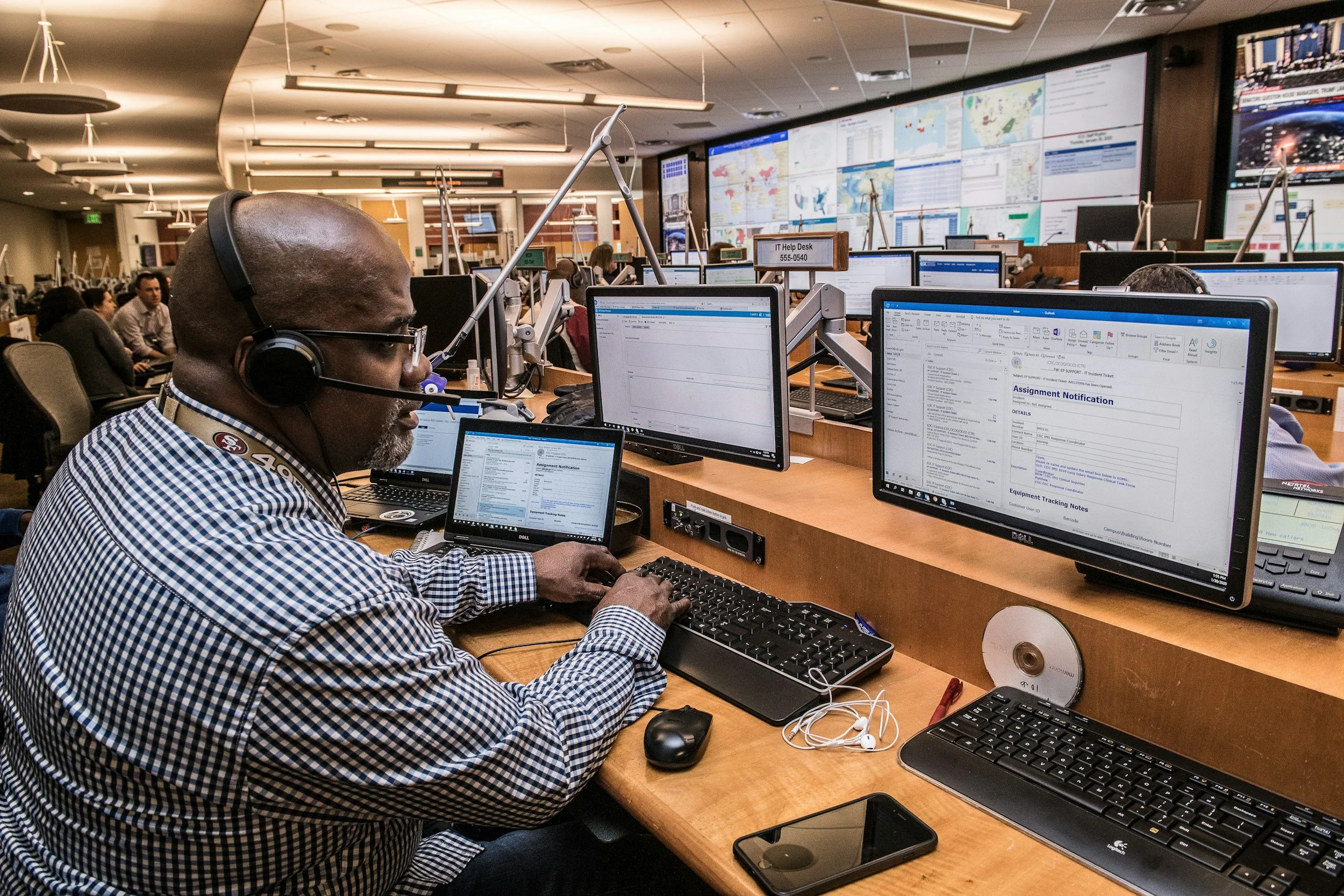 A busy workspace in a control room with multiple computer monitors, a man with a headset working at his desk, and large screens on the wall displaying maps, charts, and data.