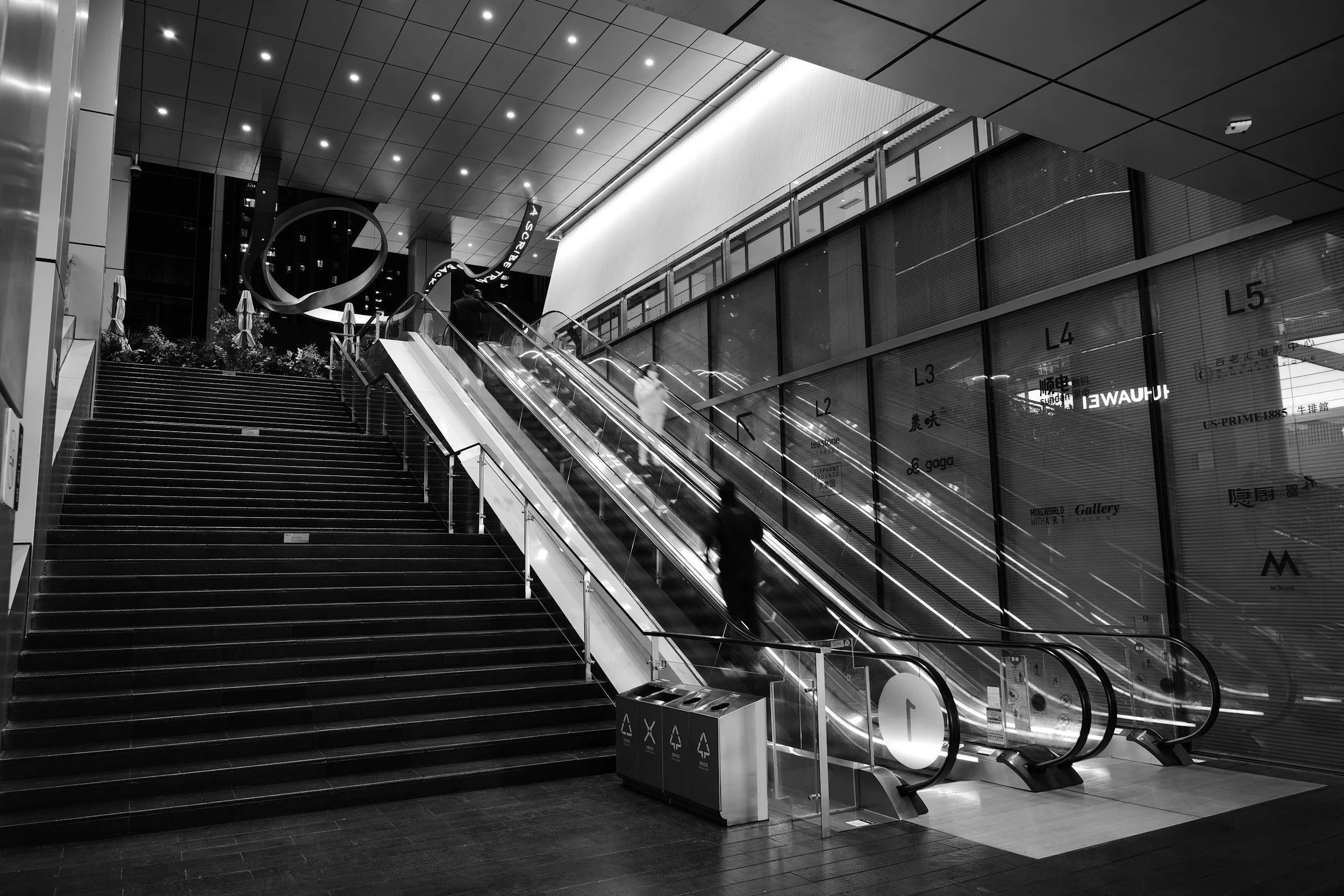 Black and white photo of an indoor shopping mall with escalators, stairs, and signage for stores. Modern architecture with artificial lighting.