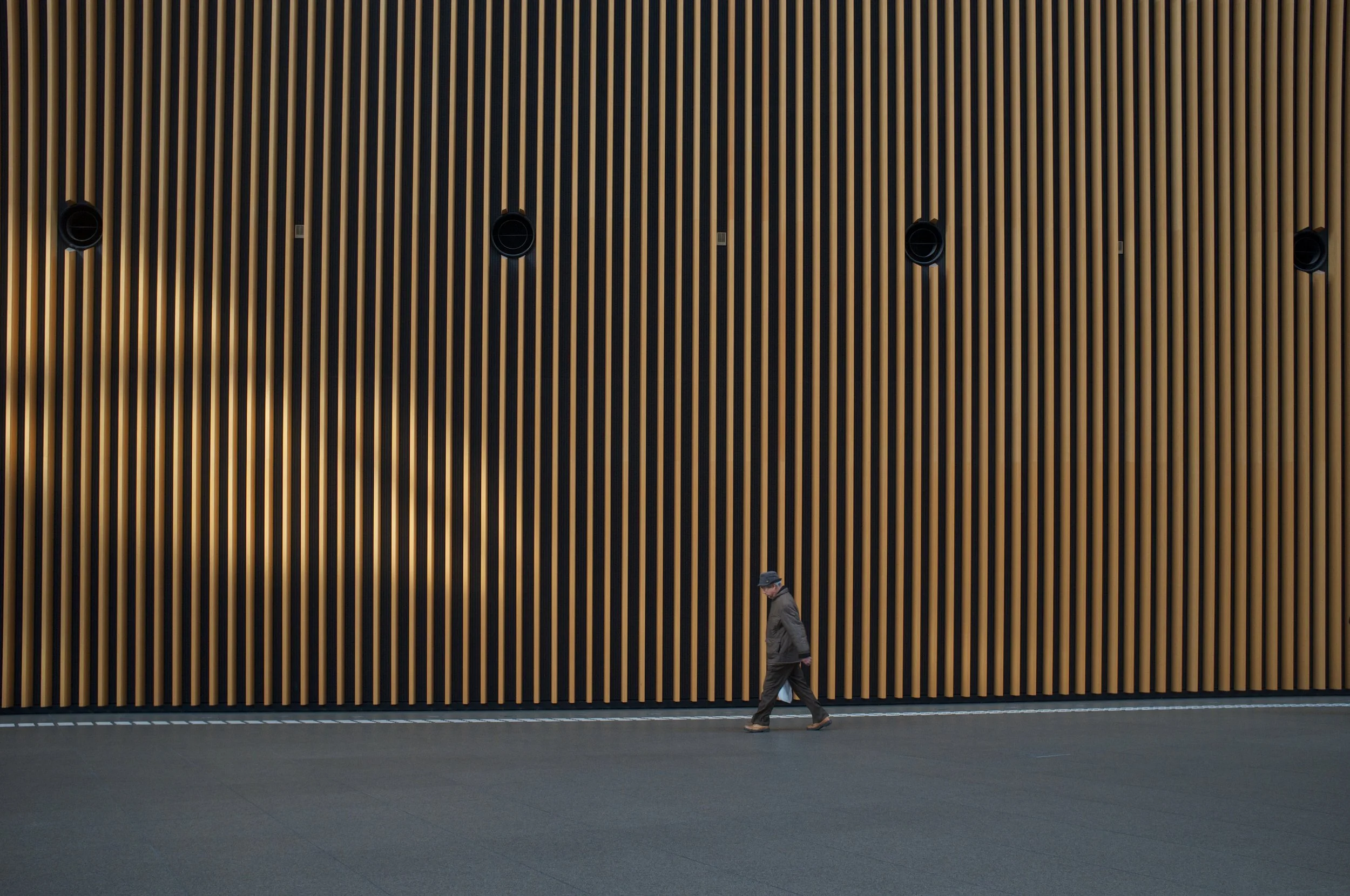 A man walking past a large wall with vertical wooden slats and built-in black circular speakers.