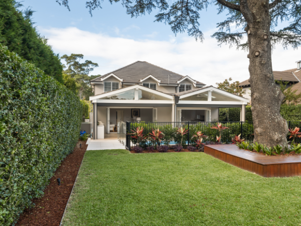 View of a backyard with a lush green lawn, hedge, flower bed, large tree, and a house with a covered patio in the background.