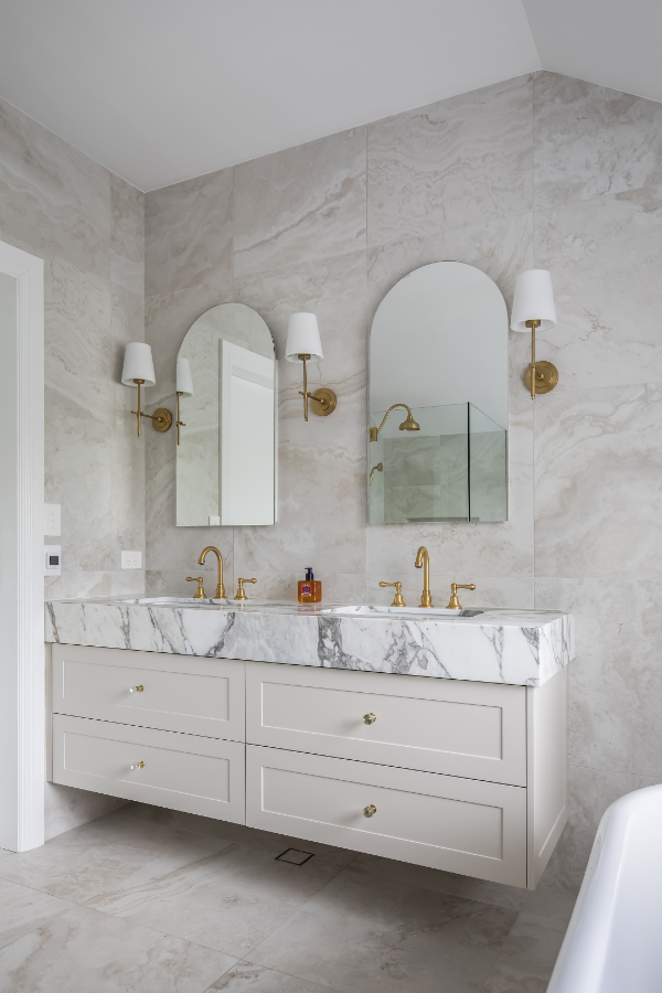 Bathroom vanity with a marble countertop, two sinks, four drawers, gold fixtures, two arched mirrors, and wall-mounted lamps.