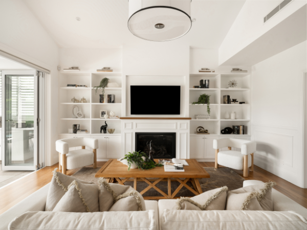 Living room with white walls, built-in white shelves, a fireplace, a large flat-screen TV, two white armchairs, a wooden coffee table with decor, a beige sofa with pillows, and a sliding glass door leading outside.