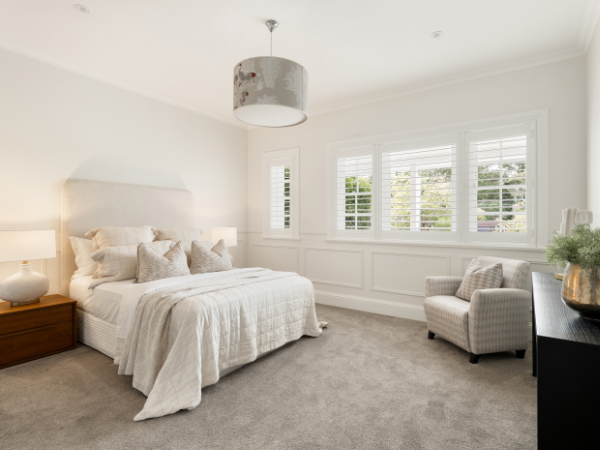 A brightly lit bedroom with a large bed, white bedding, and multiple pillows, a wooden nightstand with a lamp, a white armchair, and a set of large windows with white shutters.