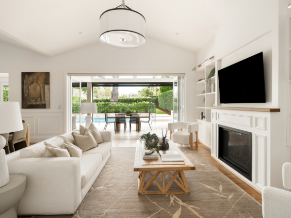 Bright living room with beige sofa, coffee table, flat-screen TV, fireplace, and large sliding glass doors opening to a backyard with greenery.