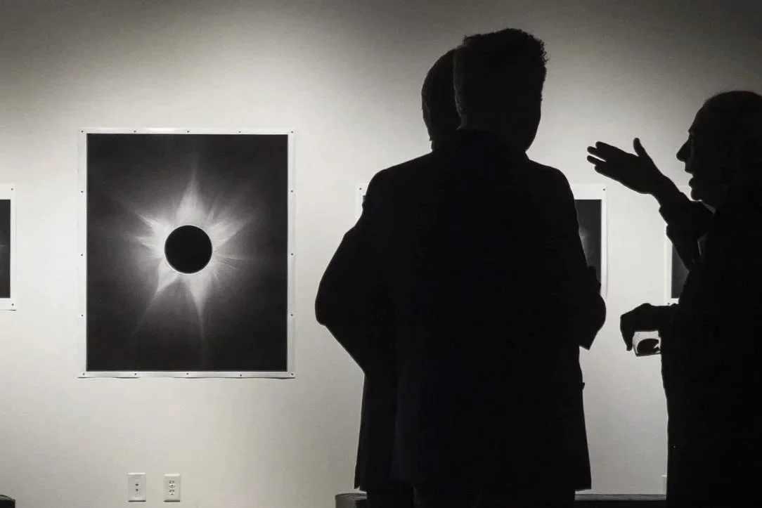 Silhouettes of three people having a conversation at an art gallery with black and white eclipse-themed photographs on the wall.