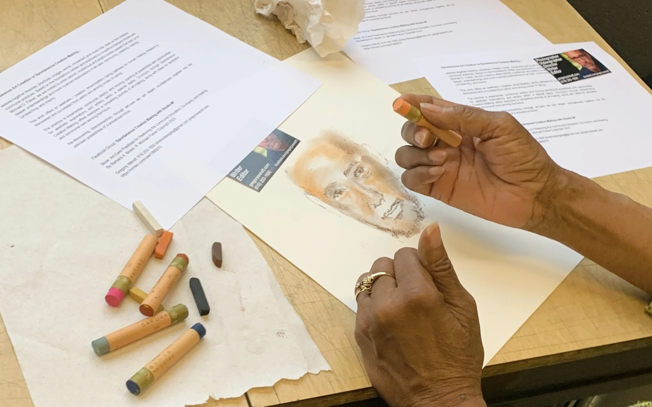 Person drawing a portrait with pastels on paper, surrounded by documents and colored chalks on a wooden table.
