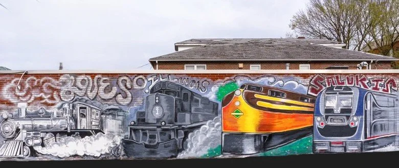 A mural painted on a brick wall depicting three trains in different styles and colors with graffiti above and to the right.