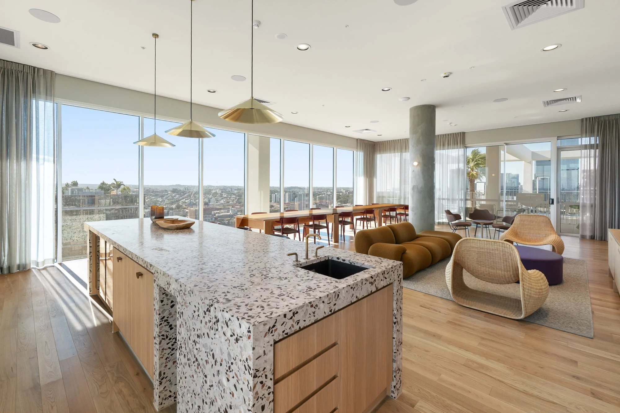 An open-concept living space with large floor-to-ceiling windows showcasing a city view, a kitchen island with terrazzo countertop, modern chairs, a brown sofa, and contemporary lighting fixtures.