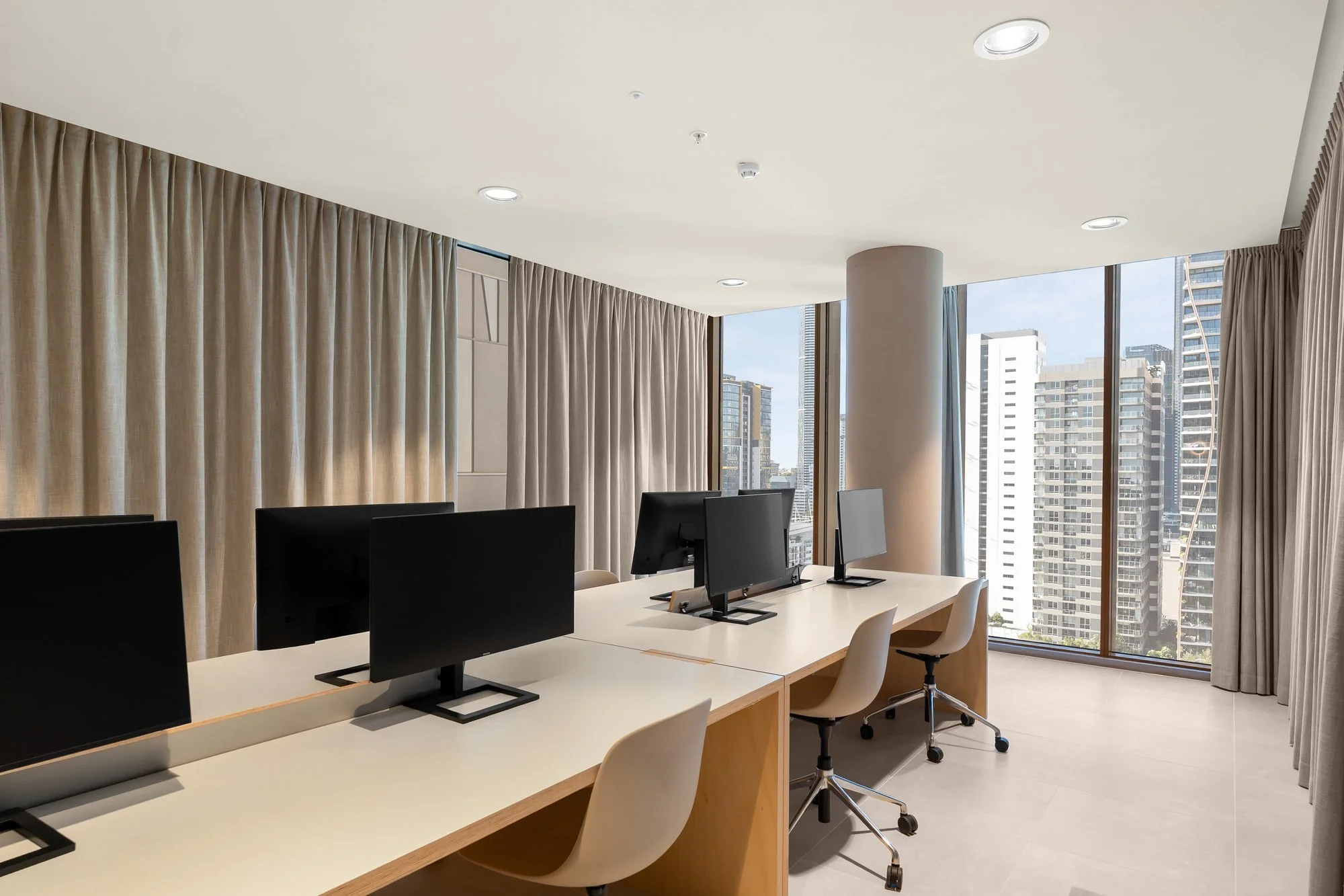 Modern office with four computer monitors on a white desk, white chairs, large windows showing city buildings, beige curtains, and ceiling lights.