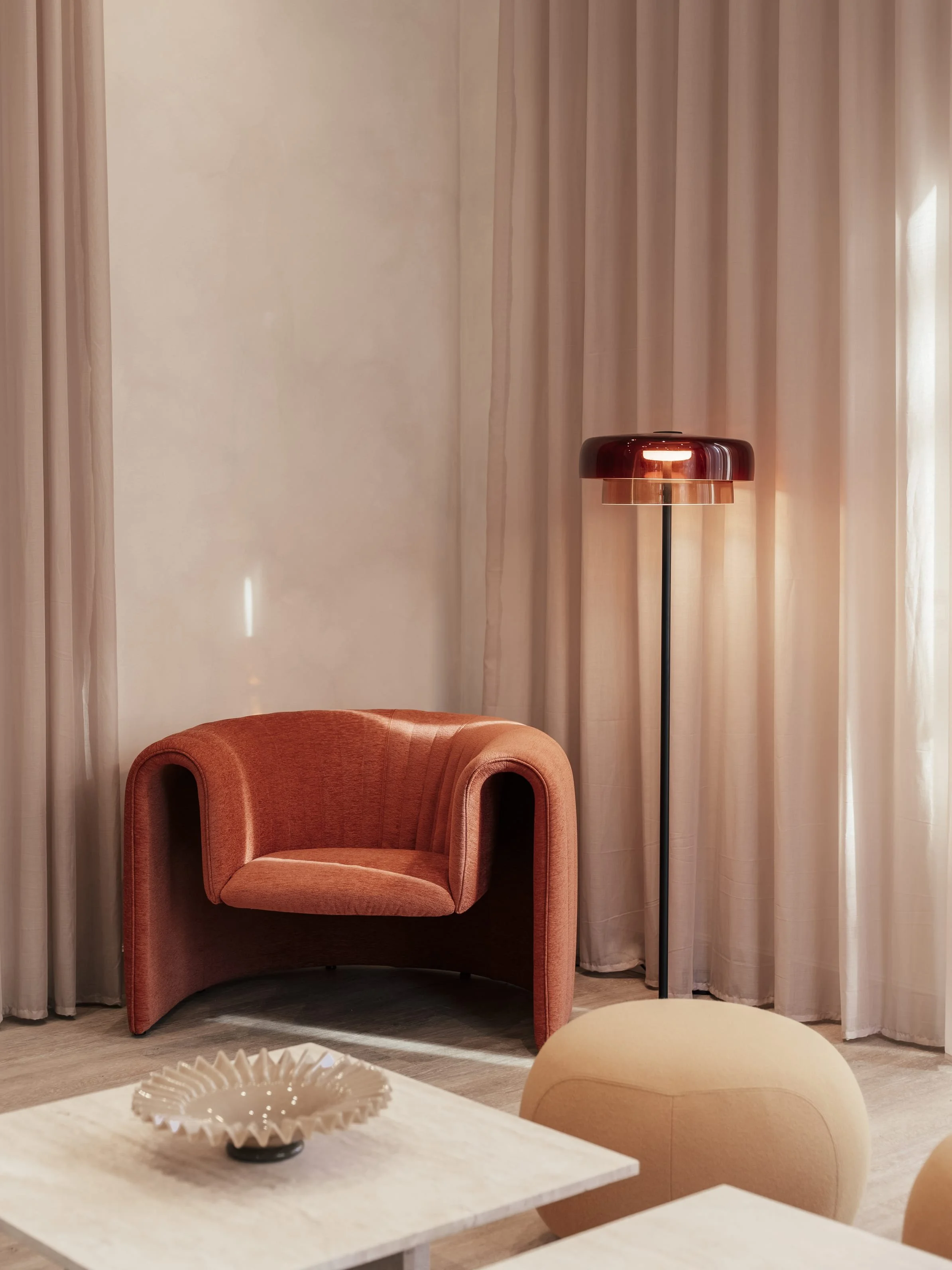 A cozy corner with a pink upholstered armchair, a tall floor lamp with a glossy dark lampshade, and cream-colored curtains in the background.