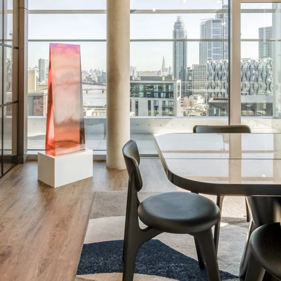 Modern office space with a view of a city skyline, a marble table, black chairs, concrete pillar, and a tall pink sculpture on a pedestal near large glass windows.