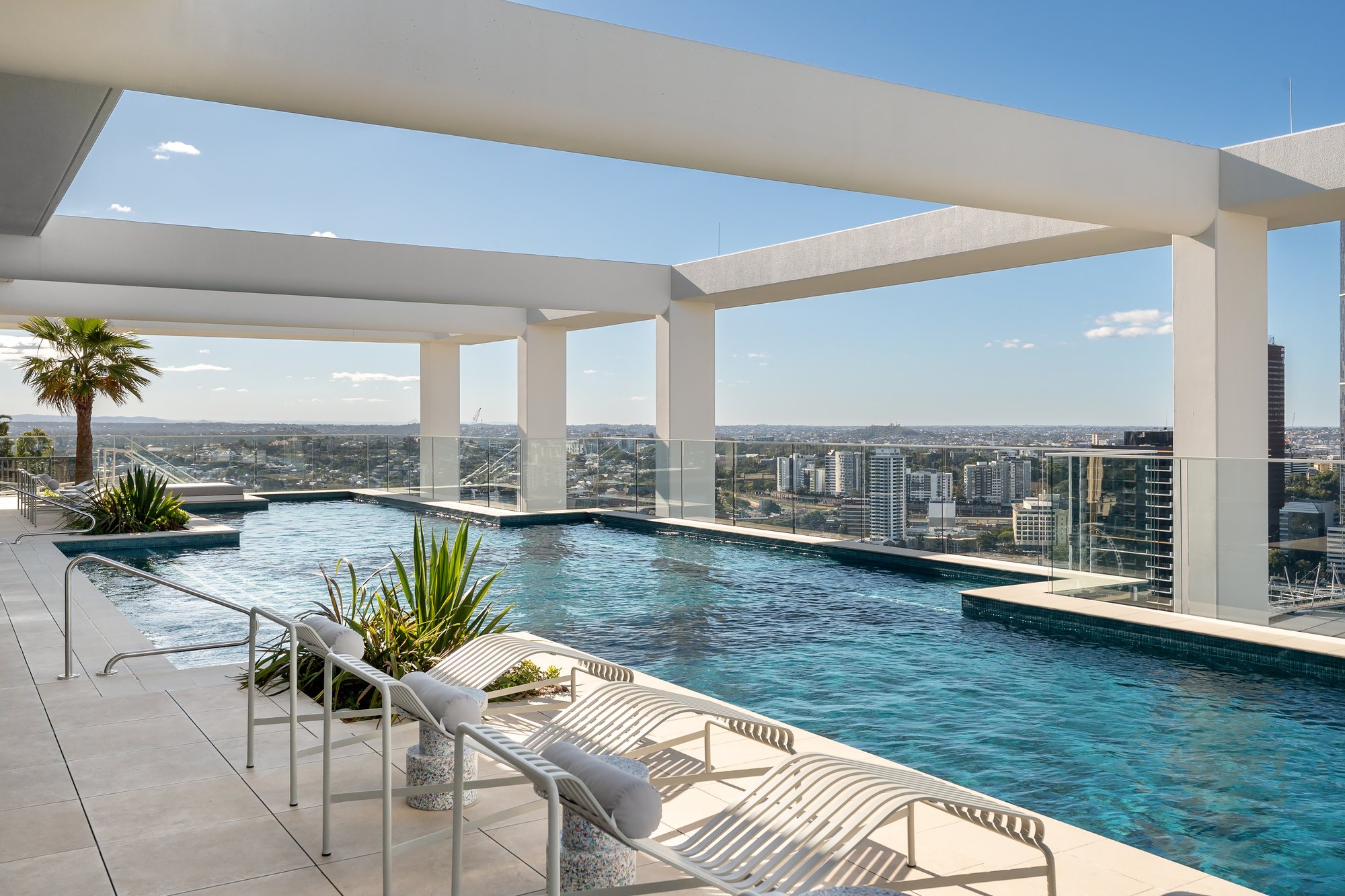 Rooftop pool with lounge chairs and potted plants under a modern white pergola overlooking a cityscape with tall buildings.