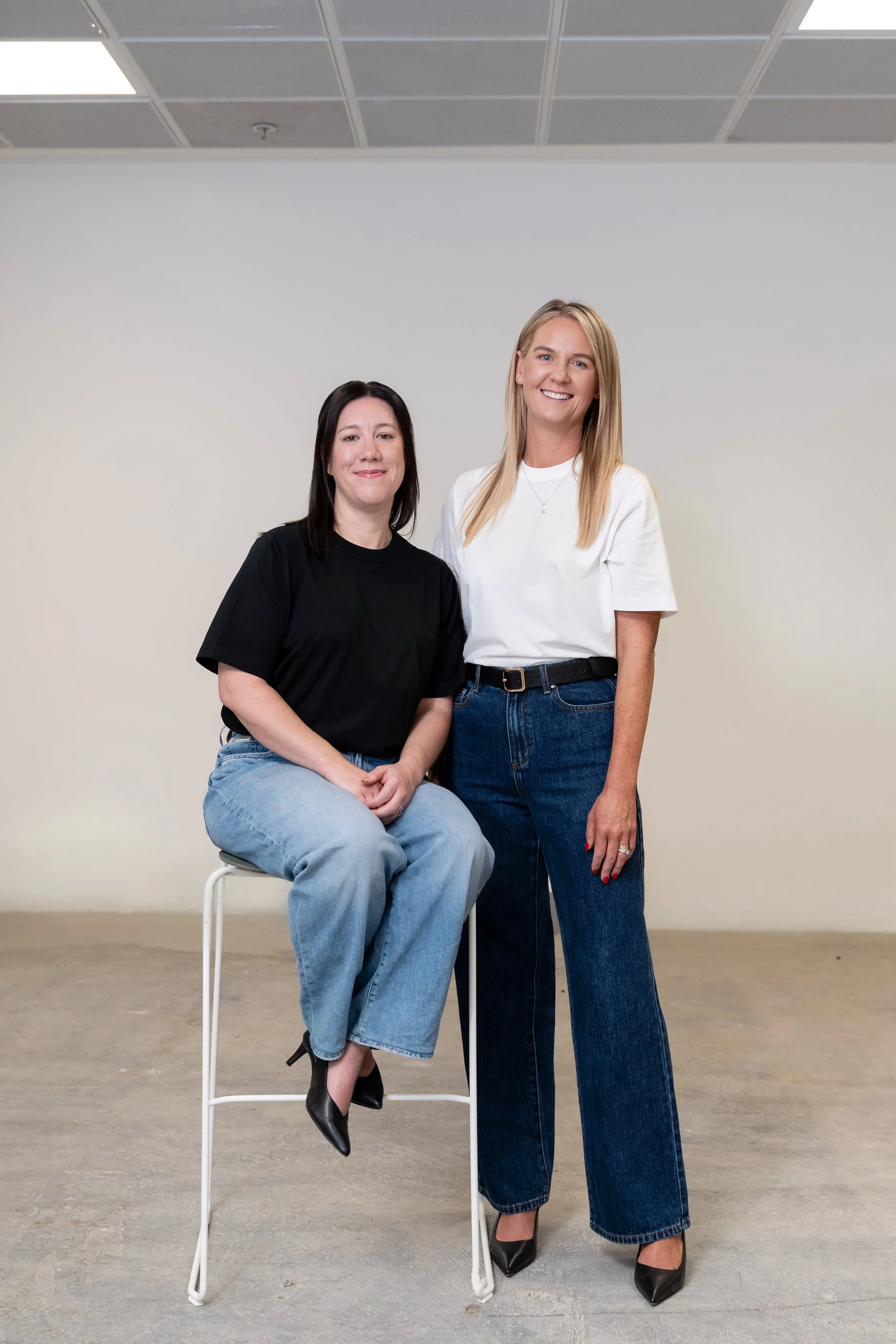 Two women, one sitting on a high stool and the other standing beside her, in an indoor setting with a plain white wall and a tiled ceiling.