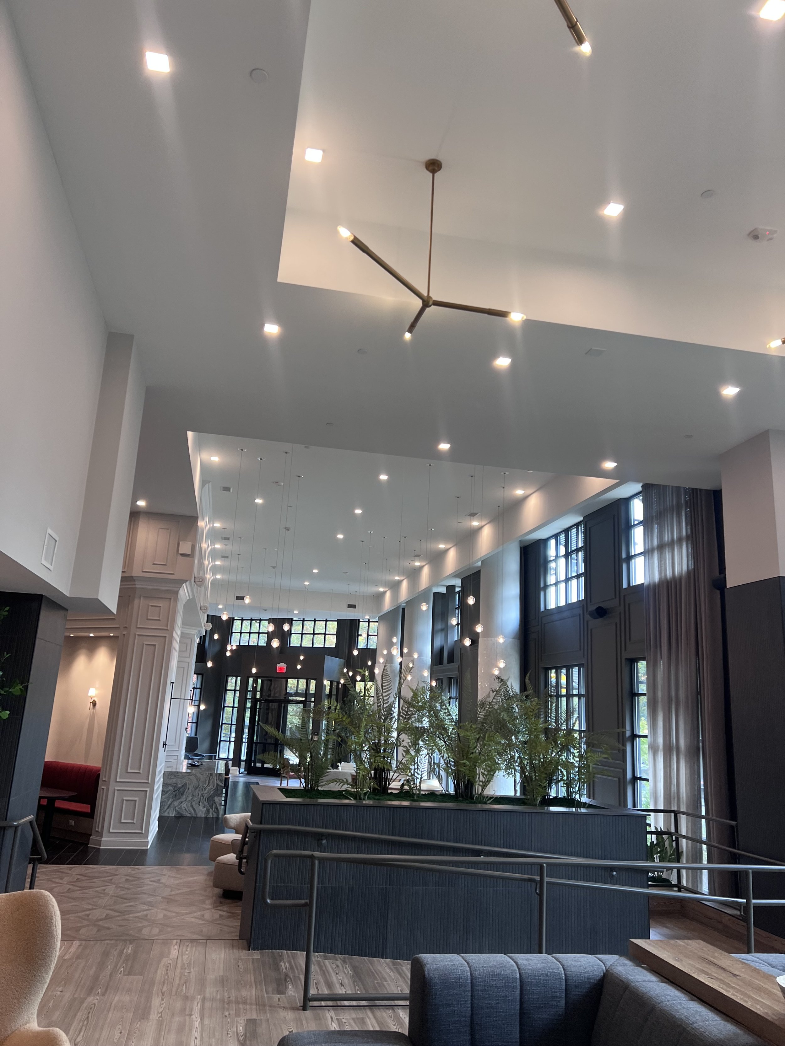 Modern hotel lobby with high ceiling, large windows, and indoor plants, furnished with chairs and sofas, illuminated by multiple ceiling lights and pendant fixtures.