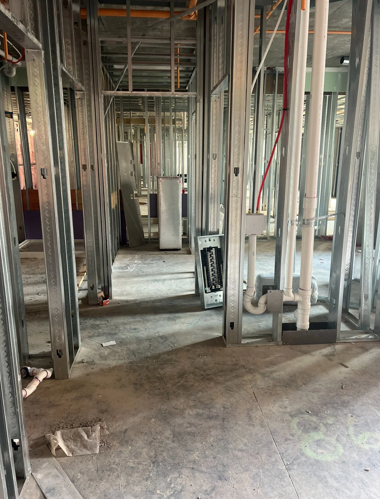 Interior view of a building under construction showing metal framing, exposed electrical and plumbing systems, and an unfinished concrete floor.