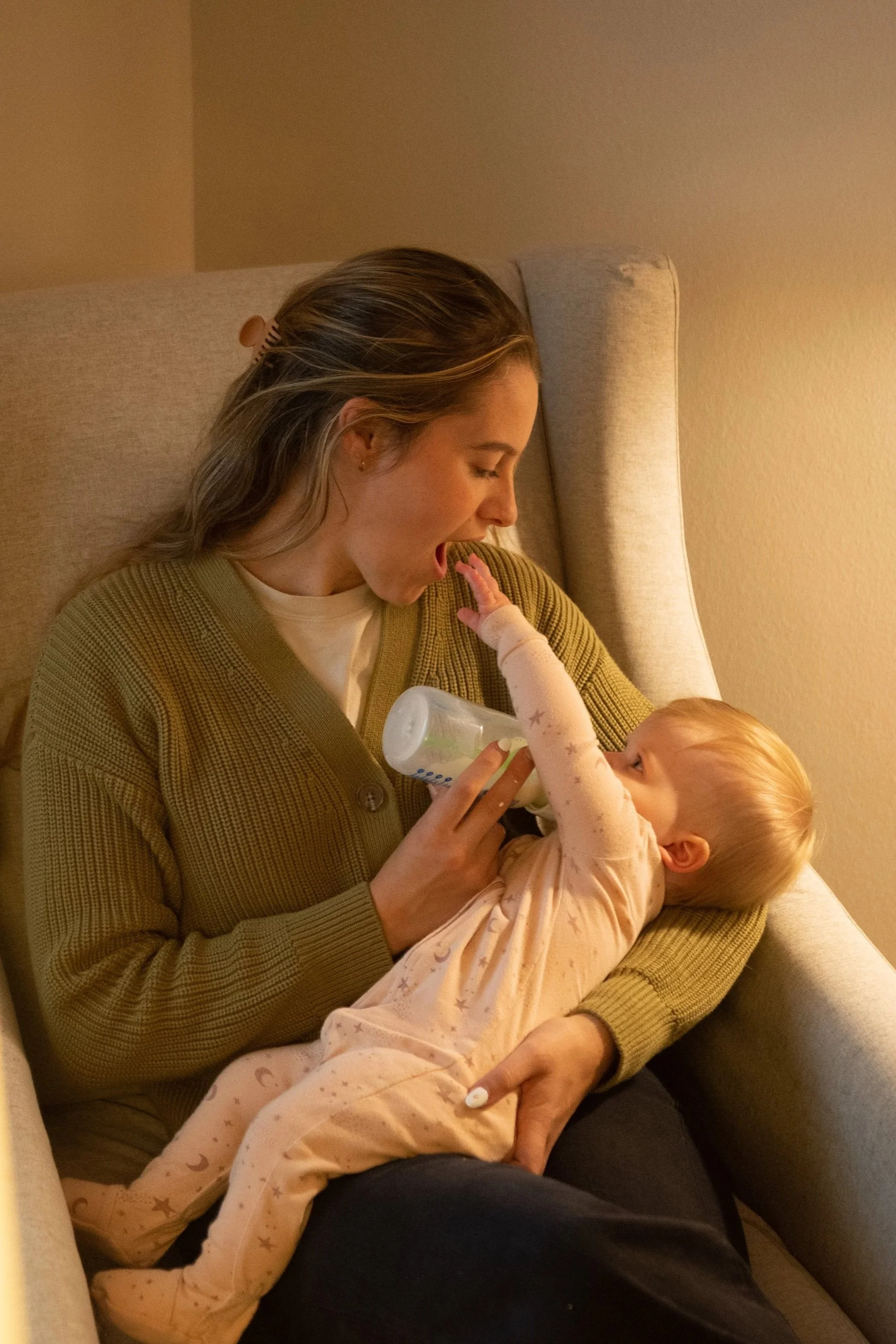 A woman in a green sweater feeding a young child with a bottle while sitting on a beige armchair.